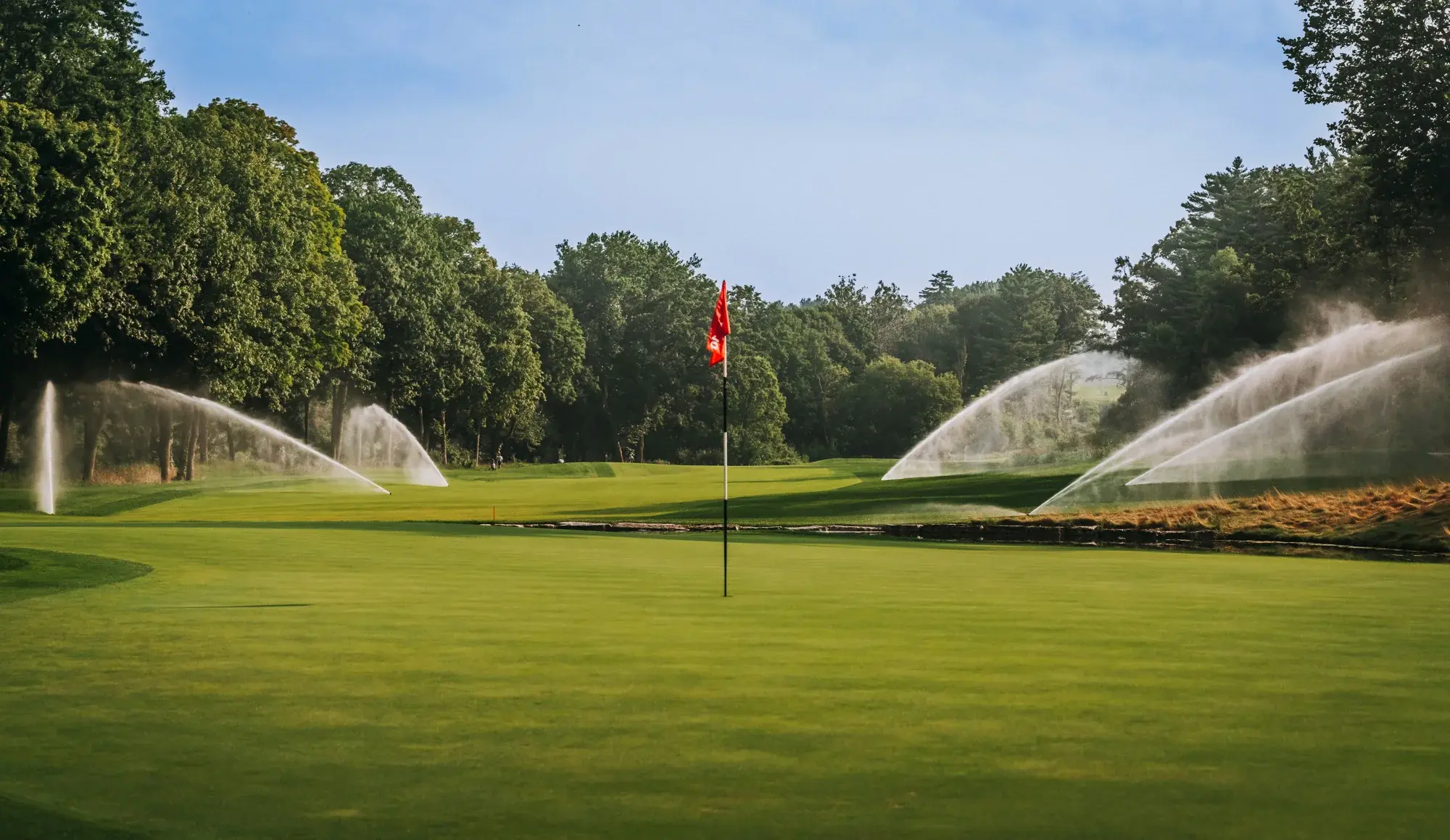 Sprinklers watering a golf green on a sunny morning, the flag up and trees behind