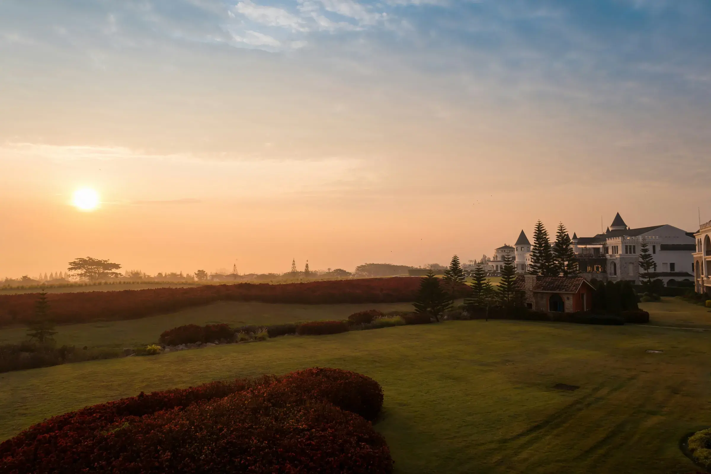 Low sun over a large estate — a clipped hedge and an empty, freshly mown lawn in the foreground, with no one on the grounds