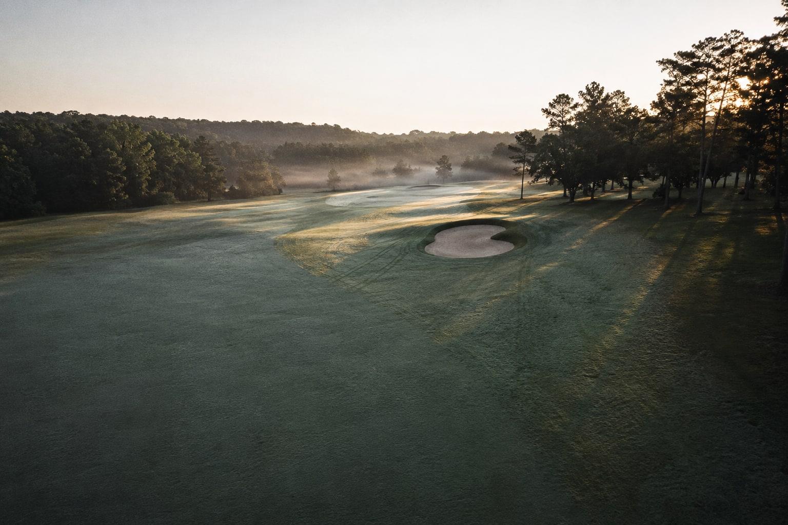 A golf course fairway at first light — mown lines still wet with dew, mist lifting off the treeline and long sun shadows across the turf, with no one on the hole