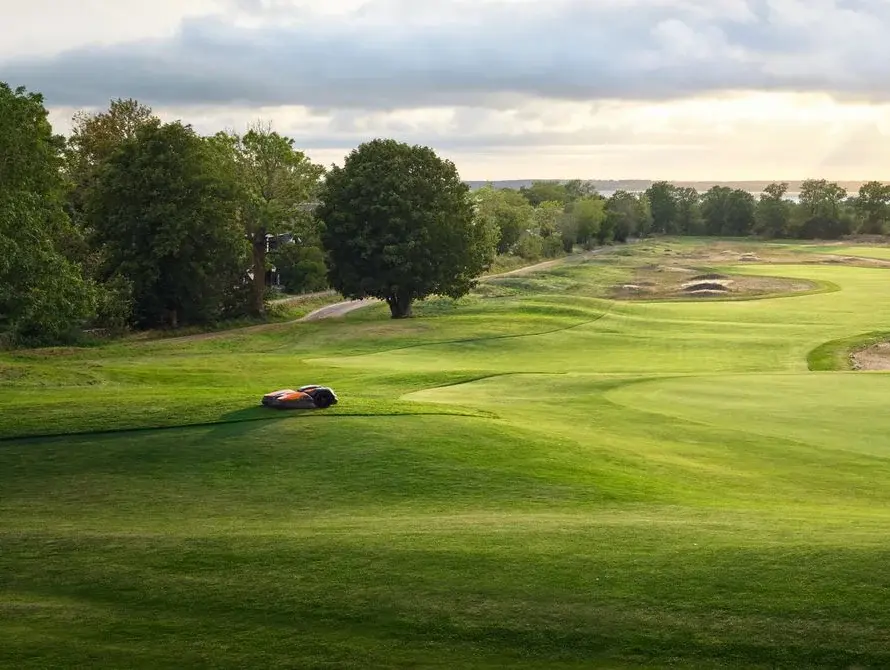 A robotic mower working the fringe of a green at golden hour while two golfers putt, the course rolling out to the sea behind