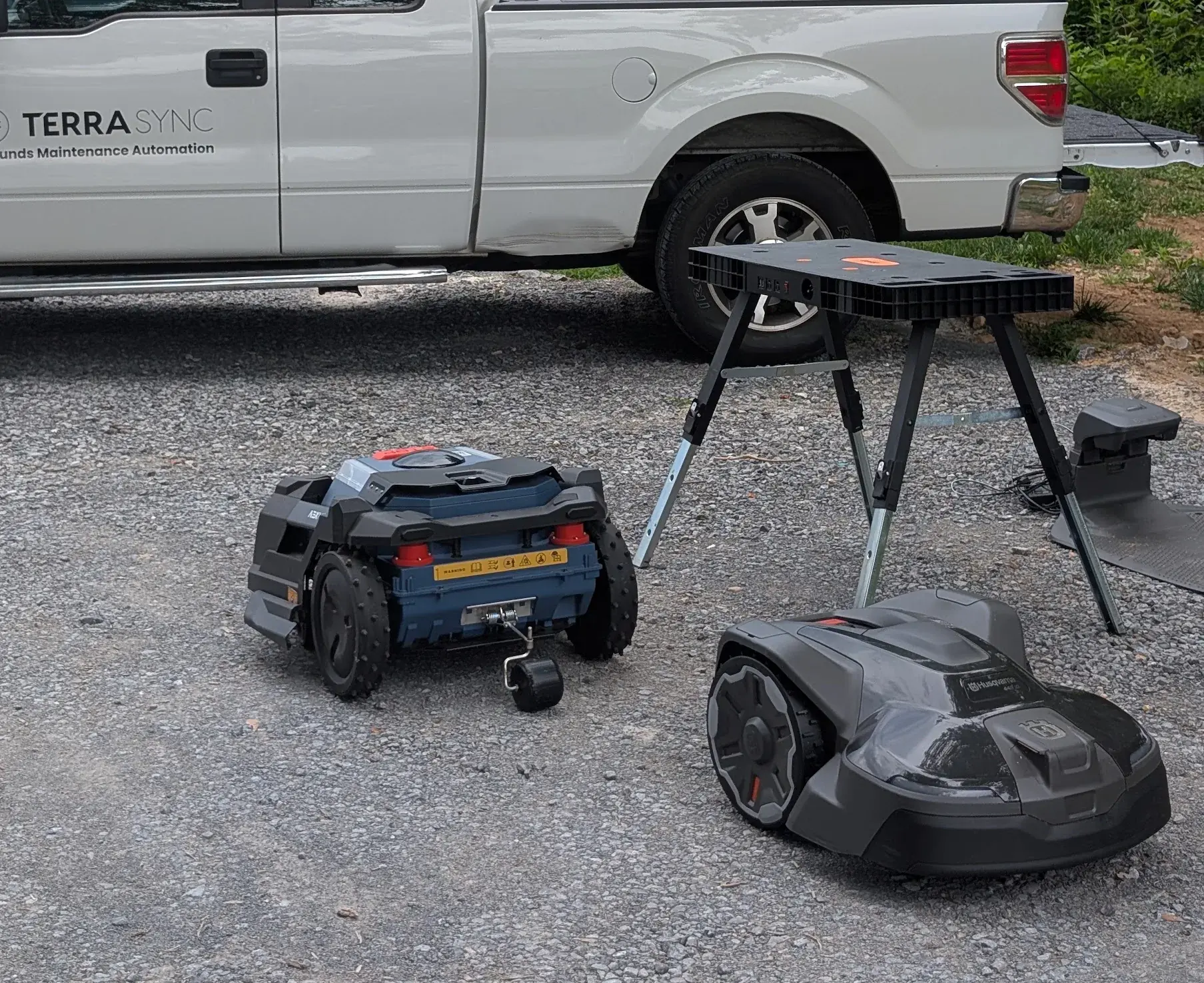 The TerraSync truck parked on gravel with a workbench and two robot mowers beside it, on a service visit