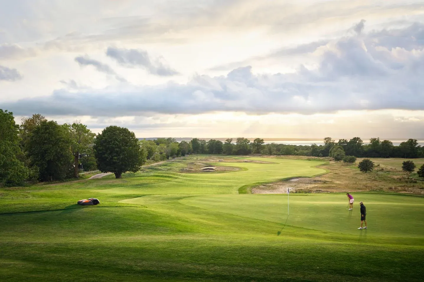 A golden-hour golf course with a robotic mower at the edge of a green while two golfers putt.